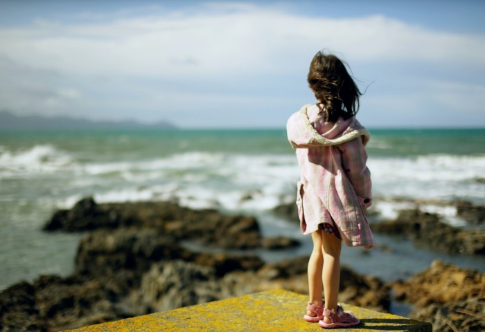 Young dark-haired girl with her back turned, overlooking a rocky seashore and out to sea.